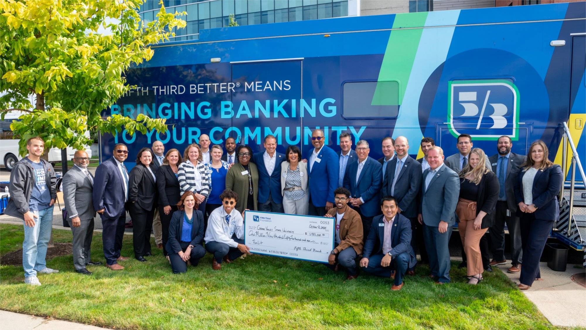 Two young men kneel in the middle and hold an oversized check made out to Grand Valley State University.